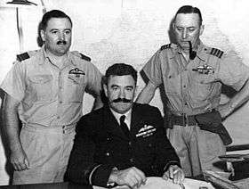 Half length portrait of three military men behind a desk, all with pilot's wings on left breast pocket. One of the men, seated, has a large dark moustache and is wearing a dark winter uniform. The other two, standing on either side of the seated figure, wear short-sleeved tropical uniforms; one of them has a small moustache, the other has a holster on his belt and is clean-shaven and smoking a pipe