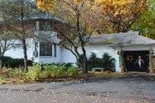  A small white brick gatehouse in front a forest of changing leaves, with a black asphalt driveway on its left