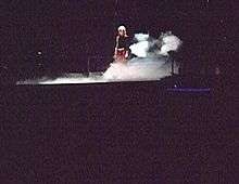 Faraway image of a woman in a short red skilt and black top standing on a stage. Smokes billow around her feet.