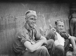 A man with a bandage around his head, smoking a cigarette beside another man with his right arm in a sling