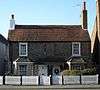 Front view of two joined flint two-storey cottages with white wooden doors next to each other under a shared tiled hood. There are two ground-floor bow windows and two tall rectangular windows above. The red-tiled roof has chimneys at each end; the right-hand one is much taller, while the left-hand one is painted white. There is a low white fence in front.