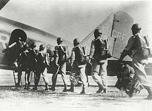 Men wearing parachutes and helmets file into a military aircraft on the ground