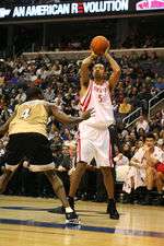 a Black athlete is shooting a basketball over another Black athlete while an Asian athlete watches.