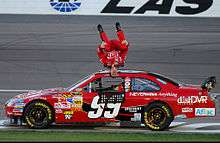 A man in his late twenties performing a backflip with the front stretch of the Las Vegas Motor Speedway in the background