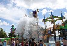 Line of people standing behind a fence looking at a colorful amusement park ride which is splashed by water.