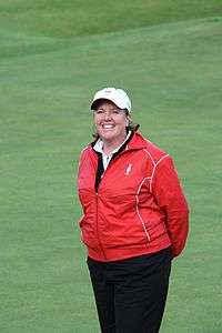 A red-haired woman in a red jacket and navy blue pants and white undershirt and white hat