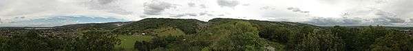 Hills covered with green vegetation. To the left in the distance are houses and water. Ahead is  exposed rock.