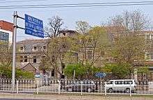 The building in the infobox and another, similar one to its right, seen from across a street in front divided by a white metal fence, with a blue road sign in Chinese and English at left. The gate of the fence is open and trees, with leaves slowly opening, are behind it.