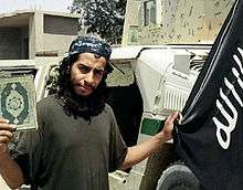 Bearded man holding a book in his right hand and a black flag in his left; a humane in the background