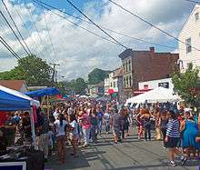 A crowded street on a nice day with tents along either side and two- to three-story buildings along the right, seen from a slight rise