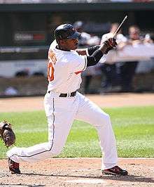 A dark-skinned man in a white baseball uniform reading "Jones" and "10" on the back in orange script swings a black baseball bat. He is wearing a black baseball helmet, and has a goatee on his chin.