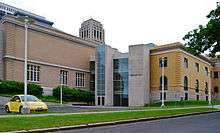 A light orange brick building with a stone and glass section connecting it to a yellow brick building, the rear of the building in the lede image. A tower rises in the distance over the structure, and there is a parking lot in front with a yellow Volkswagen New Beetle.