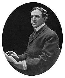 Black and white portrait photo of a white man with dark hair. He is wearing a dark suit and holding papers in his left hand.