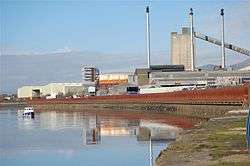 A glassworks building with large towers on the banks of an area of water