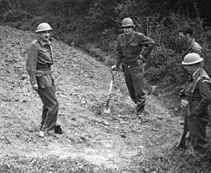 Four men in uniforms. One is leaning on a spade, and wearing an American helmet. Two others are wearing British helmets. A dark haired man has no helmet.