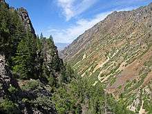 Two steep, partially wooded slopes with a distant plain visible between them. A mountain range is in the background.