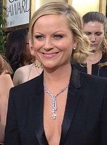 A shot from the shoulders up of a blond woman with blue eyes wearing a white and green dress, smiling and looking at something outside the image.