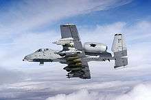 Aircraft in-flight above clouds, banking away from camera, revealing bombs and other weapons suspended underneath wings.