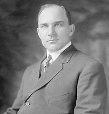 A man with receding black hair wearing a black jacket and tie and white shirt