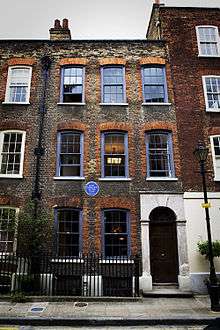 Early Georgian brick terraced house, on four levels, including the basement. Each upper floor has three sash windows. The ground floor has two and a wooden entrance door. The basement is separated from street access by spiked metal railings. A single chimney stack is on the roof, three chimney pots are visible. A blue plaque is at ground floor level. A Victorian (formally gas) street lamp is to the right of the house