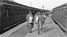 Soldiers walk down a station platform, while on either side of the platform are two trains. The men are wearing slouch hats and are carrying bags over their shoulders.