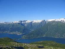 Tall, steep, snow-capped mountains line a narrow body of water. A small village sits on a narrow strip of flat ground at the base.