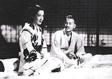 Two seated Japanese persons in traditional dress: to the left, a young woman with dark hair facing right; to the right, an elderly looking gentleman with gray hair, looking at the woman. They are sitting on futons and a shoji screen is in the background.