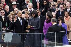 Barack Obama holds his right hand in the air as Michelle Obama looks at him and Malia and Sasha Obama watch a man whose back is to the camera looks down.