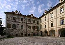 Two adjacent buildings at right angles to one another; one building has ornately painted exterior walls; the other has walls that are plainer. They have three levels of windows, and both overlook a courtyard. The background has wooded hills.