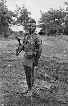 A bare-footed man with a short back and sides haircut wearing an army shirt and short skirt, carrying a weapon on a shoulder strap.