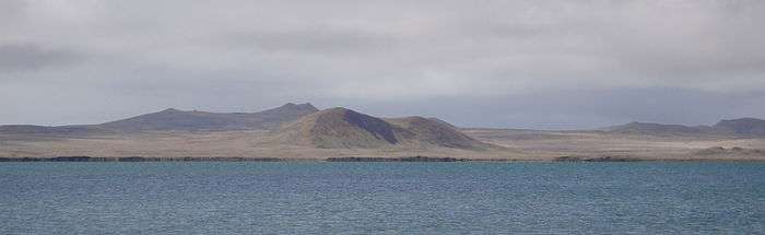 Smooth, rounded hills and flatlands covered in golden-brown vegetation lie beyond a lake under heavy cloud.