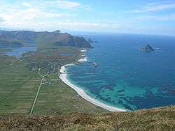 A flat, green landscape surrounded by peaks and the sea