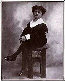 A black-and-white photograph of a curly-haired young boy, seated with one leg crossed over the other, and wearing a sailor suit.