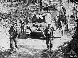 Soldiers in combat equipment carrying rifles advance along a dirt road in front of a tank