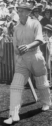 Black and white image of a man in cap and white cricket kit with batting pads and gloves on, walking onto a cricket ground. A number of spectators are in the background behind a fence.