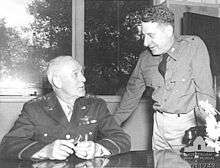 A man in uniform sits at a desk. He wears three stars and wings on his breast. Another, in a similar uniform but wearing only one star, leans over.