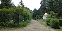 Heavily overgrown rectangular structure and a white stone structure with a gilded dome, on opposite sides of a long straight path.