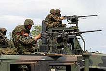 In the foreground, a HMMWV, with a MTVR in the background. Both vehicles have M2 machine guns mounted and U.S. Marines firing them.