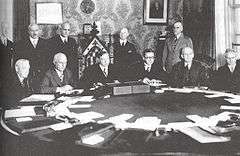 Six white men sitting around a conference table, with five more standing behind them. One of the seated men, third from the left, is sitting in a much more ornate chair than the others; he is the Prime Minister of Canada, William Lyon MacKenzie King.