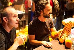 A man with short brown hair, sitting next to a man with curly black hair hugging a plush giraffe, both smiling at something to the right of the camera.