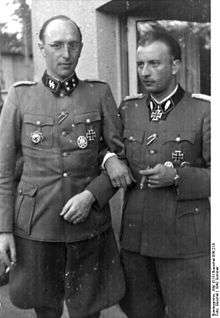 A black-and-white photograph of two men wearing military uniforms with various military decorations attached to their uniforms. The man on the left is wearing glasses and is smoking a cigarette, the man on the right is smoking a cigar.