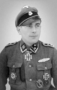 A man wearing a peaked cap with skull emblem, a military uniform with various military decorations and an Iron Cross displayed at the front of his uniform collar.