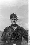 A black-and-white photograph of a man wearing a camouflage military uniform, side cap and a pair of binoculars around his neck. His cap has an emblem in shape of a human skull and crossed bones.