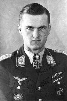 The head and shoulders of a young man, shown in semi-profile. He wears a military uniform with an Iron Cross displayed at the front of his shirt collar. His hair isshort and combed to his right, his nose is short, and his facial expression is determined; looking into the camera.