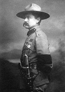 Photo of Burnham taken in 1901 in London after his investiture with the cross of the Distinguished Service Order by King Edward VII.  He is dressed in British Army uniform and standing at attention, facing right.  On his left arm is a black armband worn in mourning for the recent death of Queen Victoria. He is wearing his stetson hat and a sword on his left side. He sports a large handlebar mustache.