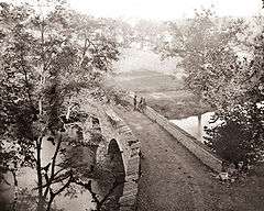 A black and white photograph of a stone bridge, viewed from above. The bridge is damaged.