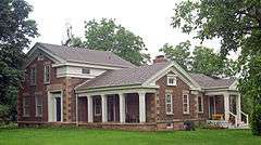 A brown house with little stones in its surface. It has two wings and some areas with square columns