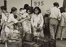 Two women stand behind their bags while a young boy and a younger woman stand to their left. A second boy is carried in a sling by the younger woman.