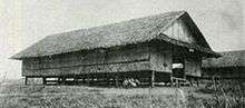 Black-and-white image a grass nipa hut raised a few feet off the ground by wooden supports. Another hut can be seen in the background.