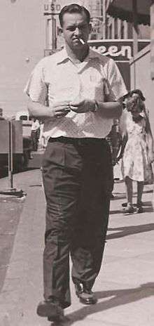 A man, with a cigarette in his mouth, is on a city sidewalk, walking and looking toward the camera.